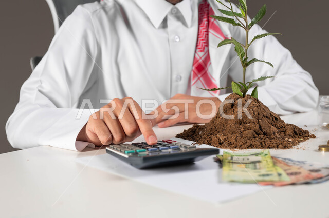 الإحصائيات والقروض البنكية, العمل في إدارة الحسابات, بيئة العمل المكتبية, بورتريه لرجل عربي خليجي سعودي يرتدي الثوب التقليدي والشماغ يقوم بإجراء العمليات الحسابية عبر الألة الحاسبة, مفهوم الوفرة المالية, التعامل بالعملات النقدية والورقية المعدنية, خلفية رمادية