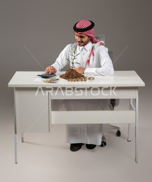 الإحصائيات والقروض البنكية, العمل في إدارة الحسابات, بيئة العمل المكتبية, بورتريه لرجل عربي خليجي سعودي يرتدي الثوب التقليدي والشماغ يقوم بإجراء العمليات الحسابية عبر الألة الحاسبة, مفهوم الوفرة المالية, التعامل بالعملات النقدية والورقية المعدنية, خلفية رمادية
