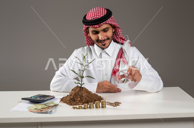 National metal and silver coins, using a calculator to postpone calculations, finance, investment and economic projects, a portrait of a smiling Saudi Gulf Arab man wearing a traditional thobe and shemagh holding a piggy bank in his hand, financial abundance and the concept of saving, gray background