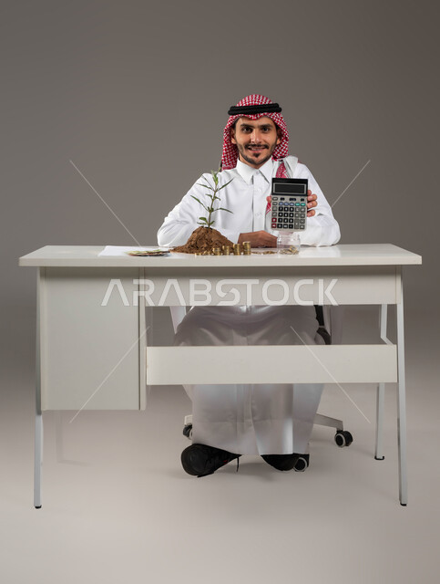الاستثمار ودراسة المشاريع الاقتصادية, إدخار الأموال والتخطيط للمستقبل, النمو والأرباح ومفهوم الوفرة المالية, بورتريه لرجل عربي خليجي سعودي يرتدي الثوب التقليدي والشماغ مبتسم يمسك بيده الألة الحاسبة لإجراء العمليات الحسابية, خلفية رمادية