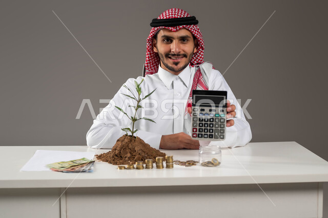 إدخار الأموال والتخطيط للمستقبل, النمو والأرباح ومفهوم الوفرة المالية, بورتريه مقرب لرجل عربي خليجي سعودي يرتدي الثوب التقليدي والشماغ مبتسم يمسك بيده الألة الحاسبة لإجراء العمليات الحسابية, الاستثمار ودراسة المشاريع الاقتصادية, خلفية رمادية