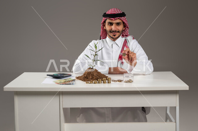 تداول الخطط للمشاريع المستقبلية, الجلوس والنظر إلى الكاميرا بإيماءات السرور والبشاشة, فوائد الإردخار المالي والاستثمار الاقتصادي, مفهوم الوفرة المالية, بورتريه لرجل عربي خليجي سعودي مبتسم يرتدي الثوب التقليدي والشماغ يمسك بيده عملة معدنية نقدية, خلفية رمادية