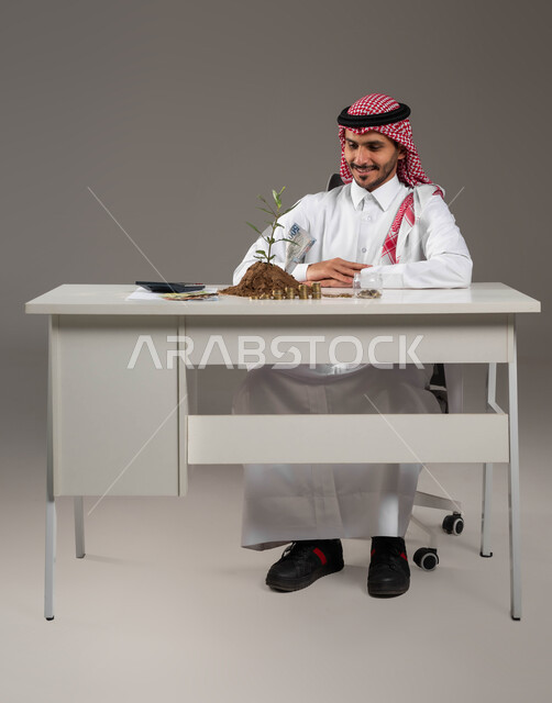 مفهوم الوفرة المالية, التخطيط في تمويل المشاريع المستقبلية, وظائف ومهن سعودية, بورتريه لرجل عربي خليجي سعودي مبتسم يرتدي الثوب التقليدي والشماغ ينظر إلى النبتة والتربة الخصبة بإيماءات السرور والسعادة بالإدخار المالي, الاستثمار في الاقتصاد السعودي, خلفية رمادية