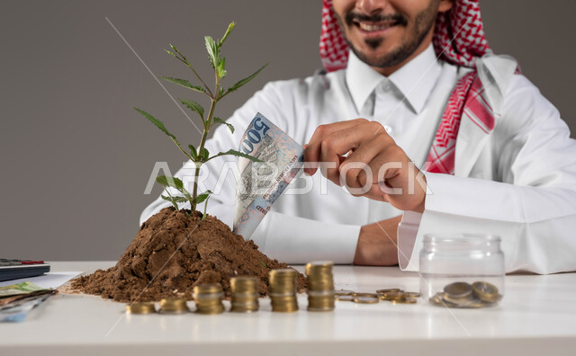 الاستثمار في القطاع الاقتصادي في المملكة, فوائد العمل والنهضة المالية, مهن وووظائف مكتبية, بورتريه لرجل عربي خليجي سعودي مبتسم يرتدي الثوب التقليدي والشماغ يمسك بيده عملة ورقية من النبتة والتربة الخصبة, مفهوم الإدخار والتوفير المالي, خلفية رمادية