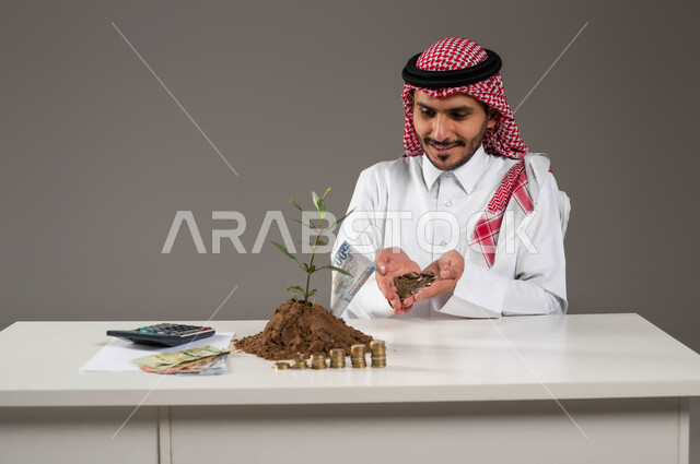مفهوم الاقتصاد والاستثمار المالي, إدخار وتوفير الأموال، بورتريه لرجل عربي خليجي سعودي مبتسم يرتدي الثوب التقليدي والشماغ يحمل بيده عملات نقدية معدنية للريال السعودي، استخدام الالة الحاسبة لإجراء العمليات الحسابية, نبتة صغيرة في تربتها الخصبة، خلفية رمادية