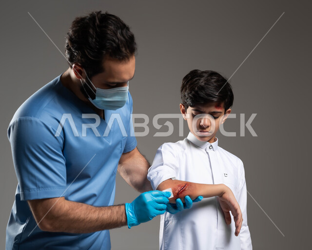 إصابات وكدمات جسدية مؤلمة، إيماءات الحزن والألم، بورتريه مقرب لممرض اسعافات اولية عربي خليجي سعودي يرتدي اليونيفورم الطبي يقوم بتعقيم الجروح لفتى، الحالات الصحية الطارئة المستعجلة، مفهوم الخدمات والرعاية الطبية المنزلية، خلفية رمادية