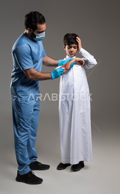 فتى سعودي يضع يده على رأسه بتعابير الألم، مفهوم الخدمات والرعاية الطبية المنزلية، الحالات الصحية الطارئة المستعجلة، بورتريه لممرض اسعافات اولية عربي خليجي سعودي يرتدي اليونيفورم الطبي يقوم بتعقيم الجروح والاصابات الجسدية للولد، خلفية رمادية