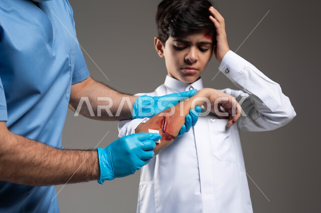 فتى سعودي يضع يده على رأسه بتعابير الألم، مفهوم الخدمات والرعاية الطبية المنزلية، الحالات الصحية الطارئة المستعجلة، بورتريه مقرب لممرض اسعافات اولية عربي خليجي سعودي يرتدي اليونيفورم الطبي يقوم بتعقيم الجروح والاصابات الجسدية للولد، خلفية رمادية