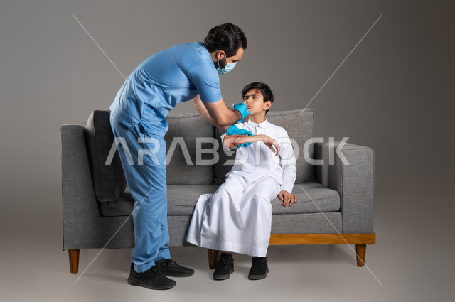 المشاكل والاصابات الجسدية، إيماءات الحزن والألم، بورتريه لممرض اسعافات اولية عربي خليجي سعودي يرتدي اليونيفورم الطبي يقوم بتعقيم الجروح لفتى على اريكة المنزل، الحالات الصحية الطارئة المستعجلة، مفهوم الخدمات والرعاية الطبية المنزلية، خلفية رمادية