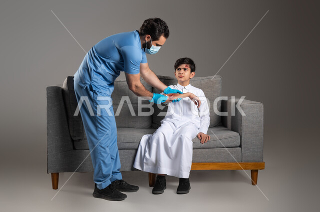 إصابات وكدمات جسدية مؤلمة، إيماءات الحزن والألم، بورتريه لممرض اسعافات اولية عربي خليجي سعودي يرتدي اليونيفورم الطبي يقوم بتعقيم الجروح لفتى على اريكة المنزل، الحالات الصحية الطارئة المستعجلة، مفهوم الخدمات والرعاية الطبية المنزلية، خلفية رمادية