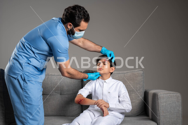 Health problems and physical injuries, portrait of a Saudi Arabian Gulf first aid nurse wearing a medical uniform sterilizing the wounds of a boy on the sofa at home, urgent emergency health cases, gestures and expressions of sadness and pain, the concept of home medical services and care, gray background