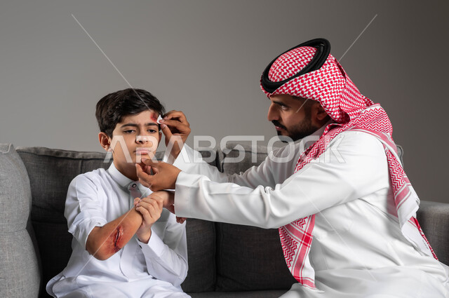اصابات وكدمات جسدية مؤلمة، تقديم الرعاية الطبية المنزلية، بورتريه مقرب لأب عربي خليجي سعودي يرتدي الشماغ والثوب التقليدي يجلس على اركية رمادية يقوم بتعقيم الجروح والالتهابات الجلدية لإبنه المصاب، مفهوم الاهتمام بصحة الأبناء، خلفية رمادية