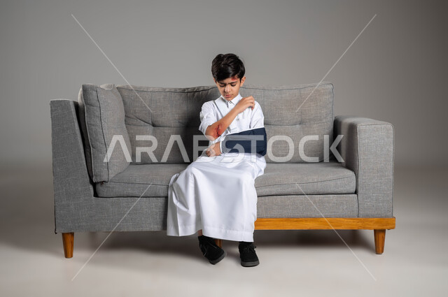 The need for home medical care, health problems and physical bruises, a portrait of a Saudi Gulf Arab boy wearing an arm sling with a broken hand sitting on the sofa, exposure to domestic violence, wounds and skin infections, expressions and gestures of pain and sadness, gray background.
