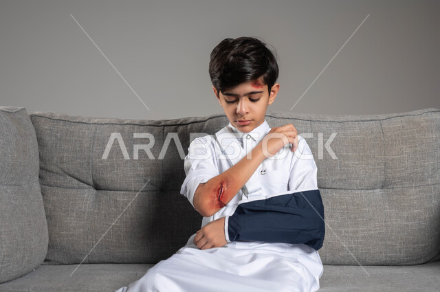 مشاكل صحية وكدمات جسدية، بورتريه مقرب لفتى عربي خليجي سعودي يرتدي حمالة الذراع بيده المكسورة يجلس على الاريكه، التعرض للعنف الأسري،  الاصابة بالجروح والالتهابات الجلدية، تعابير وايماءات الألم والحزن، الحاجة للرعاية الطبية المنزلية، خلفية رمادية