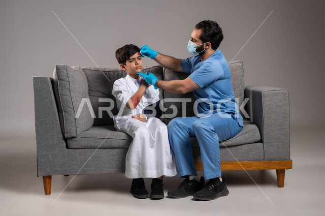 إصابات و كدمات جسدية، تعابير الألم و الحزن، المعرفة بالاسعافات الأولية للحالات الطارئة، الرعاية الصحية المنزلية، بورتريه لممرض يرتدي الكفوف والكمامة الوقائية يقوم بتعقيم جرح في جبهة ولد عربي خليجي سعودي يجلس على الكنبة، معالجة الرضوض و الكسور، خلفية رمادية