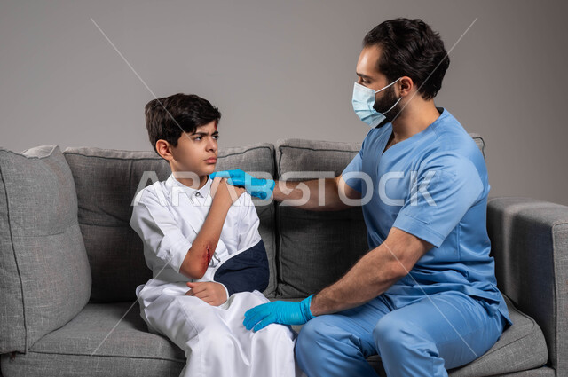 المعرفة بالاسعافات الأولية للحالات الطارئة، تقديم الاهتمام والرعاية الصحية للأبناء في المنزل، بورتريه مقرب لممرض يرتدي الكفوف والكمامة الوقائية يقوم بمواساة ولد عربي خليجي سعودي يجلس على الكنبة بإيماءات الحزن و الألم، معالجة الإصابات و الكسور، خلفية رمادية