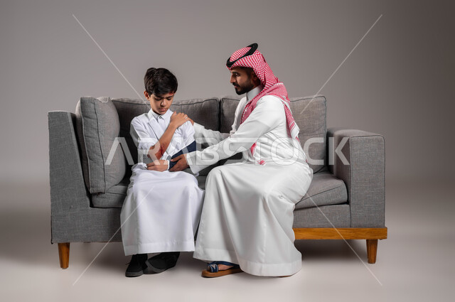 اصابات وكدمات جسدية، ارتداء الجبيرة وحمالة الذراع في اليد المكسورة، تقديم الاهتمام والرعاية الصحية للأبناء، بورتريه لأب عربي خليجي سعودي يرتدي الزي التقليدي يجلس على اريكة يقوم بمواساة إبنه المصاب، تعابير الألم والشعور بالوجع، خلفية رمادية