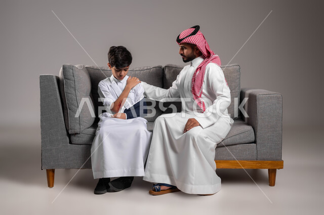 ارتداء الجبيرة وحمالة الذراع في اليد المكسورة، تقديم الاهتمام والرعاية الصحية للأبناء، بورتريه لأب عربي خليجي سعودي يرتدي الزي التقليدي يجلس على اريكة يقوم بمواساة إبنه المصاب، اصابات وكدمات جسدية، تعابير الألم والشعور بالوجع، خلفية رمادية