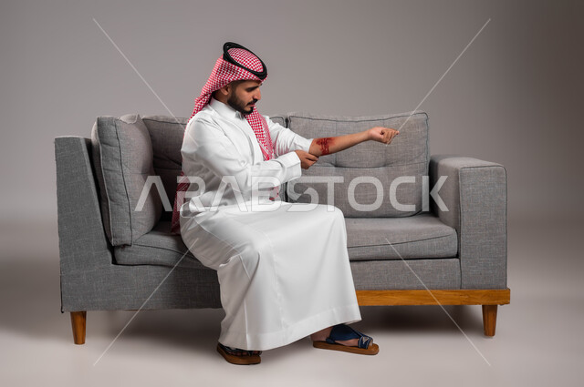 The need to visit a doctor to sterilize and bandage wounds, the necessity of training in first aid for emergency situations, a portrait of a Saudi Gulf Arab young man wearing traditional clothing sitting on a gray sofa looking at a deep wound in his arm, physical injuries and bruises, bleeding arteries and blood vessels, gray background