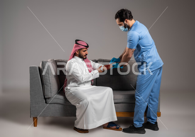 مفهوم تقديم خدمات الرعاية الصحية المنزلية، الاصابات الجسدية والمشاكل الصحية، الحالات الصحية الطارئة المستعجلة، بورتريه لممرض اسعافات اولية عربي خليجي سعودي يرتدي اليونيفورم الطبي يقوم بتعقيم الجروح لرجل على اريكة المنزل، إيماءات وتعابير الحزن والألم، خلفية رمادية
