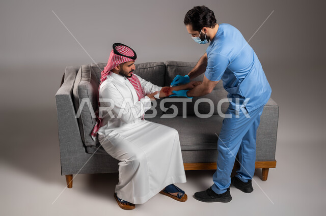 شركات الرعاية الصحية المنزلية السعوديه، المشاكل الصحية والاصابات الجسدية، بورتريه لممرض اسعافات اولية عربي خليجي سعودي يرتدي اليونيفورم الطبي يقوم بتعقيم الجروح لرجل على اريكة المنزل، الحالات الصحية الطارئة المستعجلة، إيماءات وتعابير الحزن والألم، خلفية رمادية