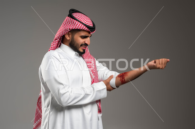 اصابات جسدية خطيرة، زيارة الطبيب لتعقيم الجروح، نزيف الشرايين والأوعية الدموية، بورتريه مقرب لشاب عربي خليجي سعودي يرتدي الزي التقليدي ينظر الى جرح عميق في ذراعه، الحاجة للرعاية الطبية المستعجلة، الاسعافات الاولية للحالات الطارئة، خلفية رمادية