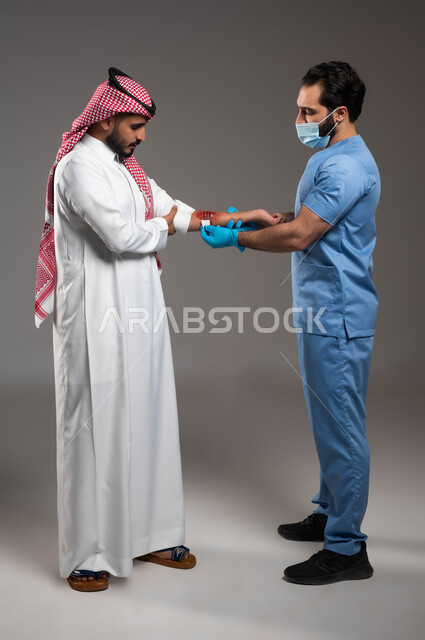 تعابير الألم والشعور بالوجع، بورتريه لممرض اسعافات اولية يرتدي اليونيفورم والكمامة الطبية يقوم بتعقيم جروح عميقة في يد رجل عربي خليجي سعودي يرتدي الشماغ والثوب التقليدي، تقديم الخدمات الطبيه والرعاية الصحية المنزلية، صورة الجسم بالكامل، خلفية رمادية