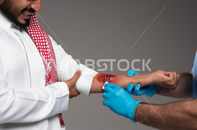إصابات وكدمات جسدية، بورتريه مقرب لممرض يرتدي القفازات الطبية يقوم بتعقيم وتطهير جروح عميقة في ذراع رجل عربي خليجي سعودي يرتدي الشماغ والثوب التقليدي، تعابير الألم والشعور بالوجع، تقديم الخدمات الطبيه والرعاية الصحية المنزلية، خلفية رمادية