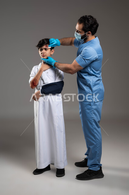 التعرض للرضوض والكسور، بورتريه لممرض اسعافات اولية عربي خليجي سعودي يرتدي اليونيفورم الطبي يقوم بتعقيم الجروح والاصابات الجسدية للولد في المنزل، مفهوم الخدمات والرعاية الطبية المنزلية، الحالات الصحية الطارئة المستعجلة، صورة لكامل الجسم، خلفية رمادية