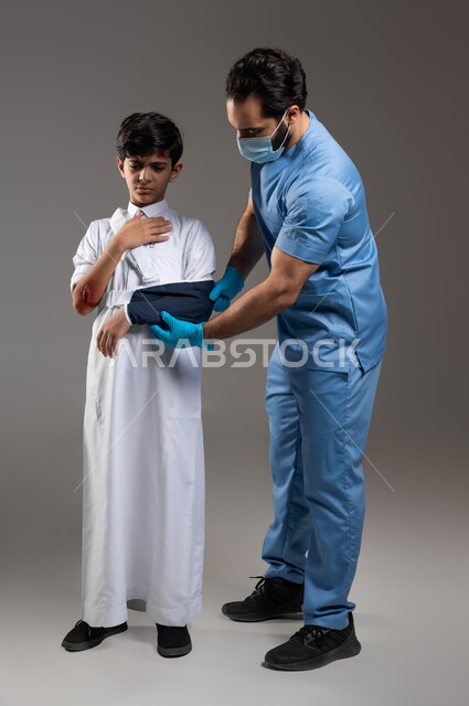 تقديم الرعاية الصحية والاسعافات الاولية المنزلية، ايماءات الألم والشعور بالوجع، بورتريه لممرض يرتدي اليونيفورم والكمامة الطبية يقوم بتركيب الجبيرة في الذراع المكسورة لولد عربي خليجي سعودي مصاب، التعرض للجروح والكسور، صورة لكامل الجسم، خلفية رمادية
