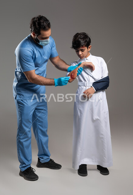 تقديم الرعاية الصحية والاسعافات الاولية المنزلية، ايماءات الألم والشعور بالوجع، بورتريه لممرض يرتدي اليونيفورم والكمامة الطبية يقوم بتعقيم وتطهير جروح عميقة في ذراع ولد عربي خليجي سعودي مصاب، التعرض للكسور والرضوض والإصابات، صورة لكامل الجسم، خلفية رمادية