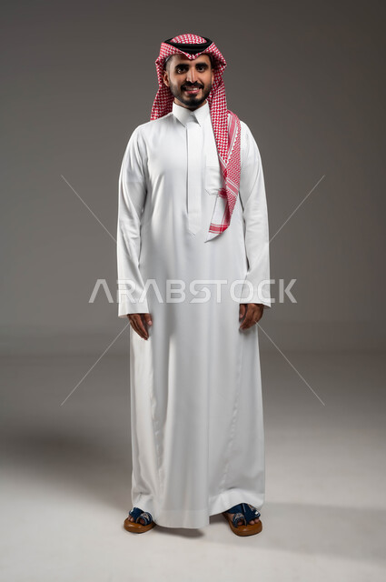 الوقوف باستقامة وانضباط، إيماءات الثقة والاعتزاز بالنفس، الاهتمام بالمظهر الخارجي، بورتريه لشاب عربي خليجي سعودي مبتسم يرتدي الشماغ والثوب التقليدي ينظر إلى الكاميرا بتعابير الجدية، مفهوم الاناقه والرجولة، صورة الجسم بالطول الكامل، خلفية رمادية