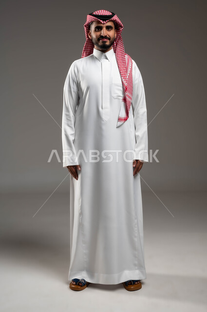 Gestures of confidence and self-esteem, attention to external appearance, portrait of a smiling Saudi Arabian Gulf man wearing a shemagh and traditional dress looking at the camera with serious expressions, the concept of elegance and masculinity, standing upright and disciplined, full-length body portrait, gray background