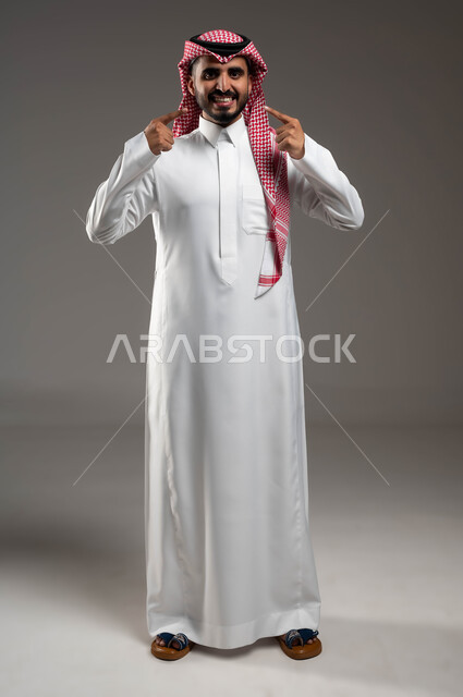 الزيارة الدورية عند الطبيب، إبتسامة هوليود باستخدام قشور فينير من الخزف، بورتريه لرجل عربي خليجي سعودي مبتسم يرتدي الشماغ والثوب التقليدي ينظر إلى الكاميرا ويشير بإصبع السبابة على أسنانه، صورة الجسم بالطول الكامل , خلفية رمادية .