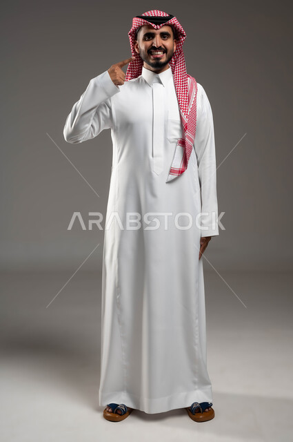 Care and attention to oral health, a Hollywood smile using porcelain veneers, a portrait of a smiling Saudi Gulf Arab man wearing a shemagh and traditional dress, looking at the camera and pointing with his index finger over his teeth, full-length body photo, gray background