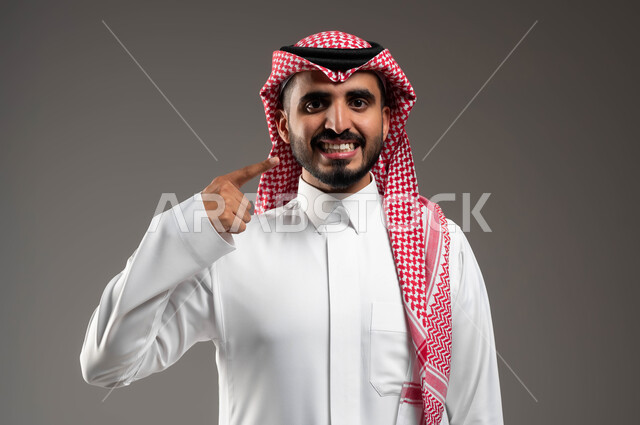 A bright white smile, gestures of joy and happiness, the use of porcelain veneers, care, beauty and whitening, a close-up portrait of a smiling Saudi Gulf Arab man wearing a shemagh and traditional thobe, looking at the camera and pointing with his index finger at his teeth, gray background