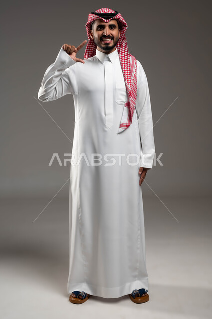 إبتسامة هوليود باستخدام قشور فينير من الخزف، بورتريه لرجل عربي خليجي سعودي مبتسم يرتدي الشماغ والثوب التقليدي ينظر إلى الكاميرا ويشير بإيماءات التحجيم، المعالجة والمحافظة على الصحة الفموية, صورة الجسم بالطول الكامل, خلفية رمادية .