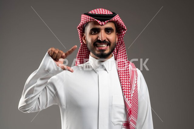 إيماءات الفرح والسعادة، استخدام قشور فينير من الخزف، العناية والتجميل والتبييض، بورتريه مقرب لرجل عربي خليجي سعودي مبتسم يرتدي الشماغ والثوب التقليدي ينظر إلى الكاميرا ويشير بإيماءات التحجيم, ابتسامة بيضاء ناصعه، خلفية رمادية .