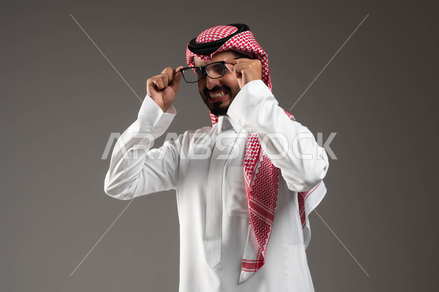 مفهوم الثقة بالنفس والرجولة، الاهتمام بالأناقة والمظهر الخارجي، بورتريه مقرب لرجل عربي خليجي سعودي مبتسم يرتدي الشماغ و الثوب التقليدي يمسك النظارة الطبية بإيماءات التركيز والتمعن والتدقيق بموضوع ما, الشعور بالفرح والسرور، خلفية رمادية .