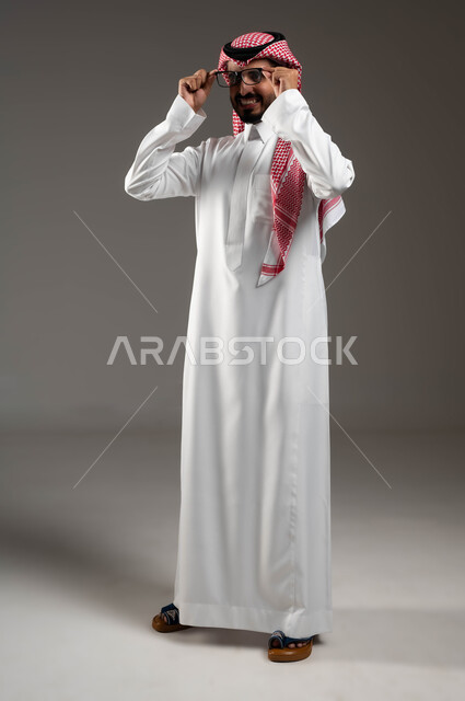 الأناقة والاهتمام بالمظهر الخارجي، مفهوم الثقة بالنفس والرجولة، بورتريه لرجل عربي خليجي سعودي مبتسم يرتدي الشماغ و الثوب التقليدي يمسك النظارة الطبية بإيماءات التركيز والتمعن والتدقيق بموضوع ما, صورة الجسم بالطول الكامل، خلفية رمادية .