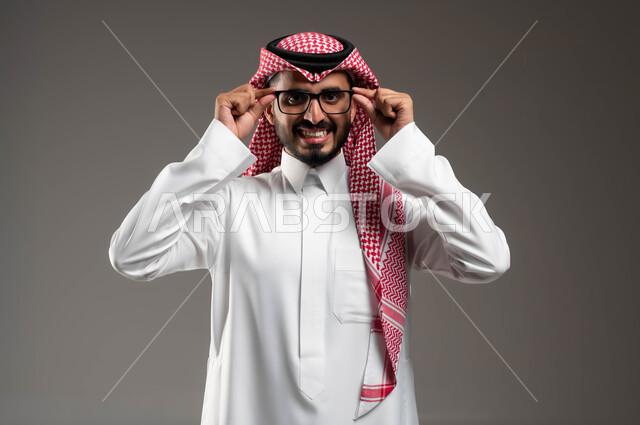 مفهوم الرجولة والثقة بالنفس، الاهتمام بالأناقة والمظهر الخارجي، بورتريه مقرب لرجل عربي خليجي سعودي مبتسم يرتدي الشماغ و الثوب التقليدي يمسك النظارة الطبية ينظر إلى الكاميرا بإيماءات التركيز, التمعن والتدقيق بموضوع ما, خلفية رمادية .