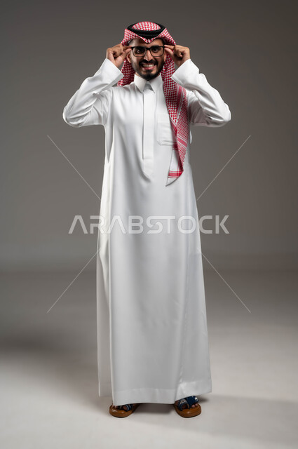 الاهتمام بالأناقة والمظهر الخارجي، الشعور بالسعادة والفرح, بورتريه لشاب عربي خليجي سعودي مبتسم يرتدي الشماغ و الثوب التقليدي يمسك النظارة الطبية ينظر إلى الكاميرا بإيماءات التركيز, التمعن والتدقيق بموضوع ما, صورة الجسم بالطول الكامل، خلفية رمادية .