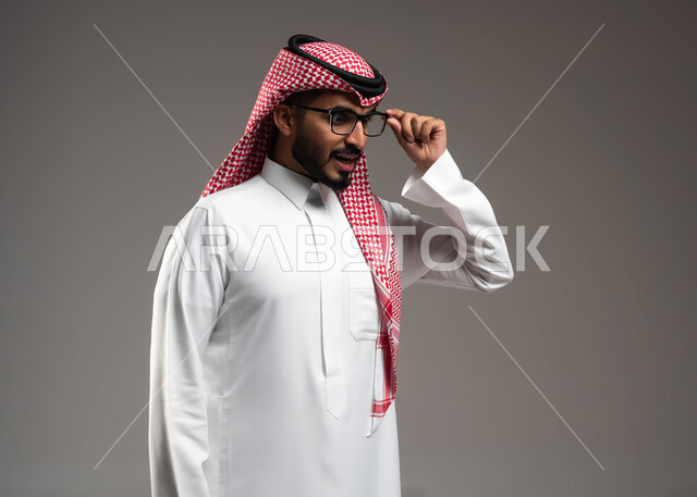 التحديق بنظرات يملؤها الذكاء والفطنة، بورتريه مقرب من الجانب لشاب عربي خليجي سعودي يرتدي الشماغ و الثوب التقليدي يمسك النظارة الطبية ينظر إلى شيء ما في الأسفل بإيماءات التركيز, خلفية رمادية .