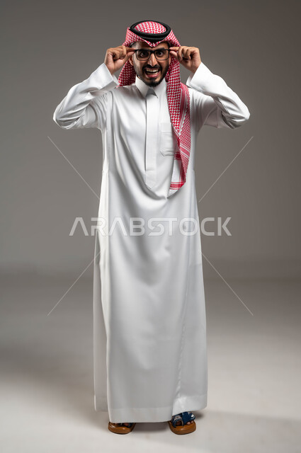 التحديق بنظرات يملؤها الذكاء والفطنة، تعابير الفرح والسرور, الشعور بالدهشة والذهول, بورتريه لشاب عربي خليجي سعودي يرتدي الشماغ و الثوب التقليدي يمسك النظارة الطبية ينظر إلى الكاميرا بإيماءات التركيز, صورة الجسم بالطول الكامل، خلفية رمادية .