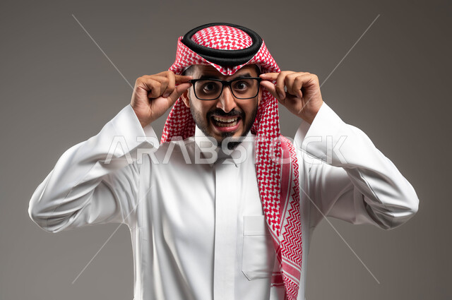 تعابير الفرح والسرور, الشعور بالدهشة والذهول, بورتريه مقرب لشاب عربي خليجي سعودي يرتدي الشماغ و الثوب التقليدي يمسك النظارة الطبية ينظر إلى الكاميرا بإيماءات التركيز, التحديق بنظرات يملؤها الذكاء والفطنة، خلفية رمادية .
