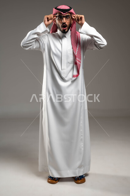 الشعور بالدهشة والذهول , بورتريه لشاب عربي خليجي سعودي يرتدي الشماغ و الثوب التقليدي يمسك النظارة الطبية ينظر إلى الكاميرا بإيماءات التركيز, الصدمة بسماع خبر ما, الاهتمام بالأناقة والمظهر الخارجي، صورة الجسم بالطول الكامل، خلفية رمادية .