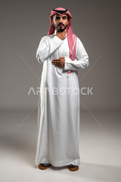 إيماءات التعب والألم وطلب المساعدة، التهاب اللوز والحاجة للمضادات الحيوية، بورتريه لرجل عربي سعودي خليجي يرتدي الشماغ والثوب التقليدي ينظر إلى الكاميرا يضع يده على حنجرته ويقوم بالسعال, صورة الجسم بالطول الكامل، خلفية رمادية