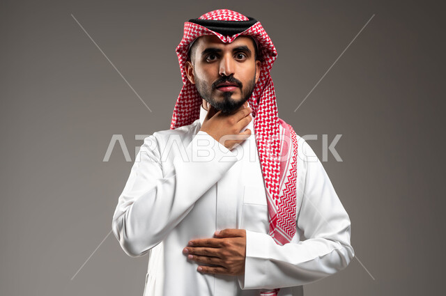 التهاب اللوز والحاجة للمضادات الحيوية، بورتريه مقرب لرجل عربي سعودي خليجي يرتدي الشماغ والثوب التقليدي ينظر إلى الكاميرا ويضع يده على حنجرته بإيماءات التعب والألم وطلب المساعدة، خلفية رمادية.