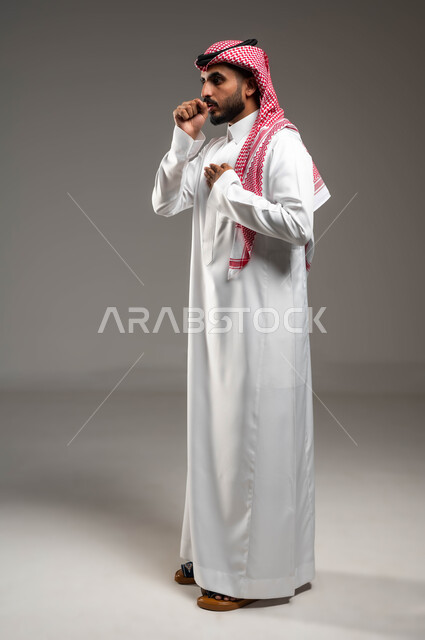 الإصابة بالأنفلونزا ونزلات البرد، الكحة والتهاب الحلق والحاجة للعلاج، بورتريه مقرب من الجانب لرجل عربي سعودي خليجي يرتدي الشماغ والثوب التقليدي ينظر إلى الكاميرا يضع يده أمام فمه ويقوم بالسعال, صورة الجسم بالطول الكامل، خلفية رمادية .