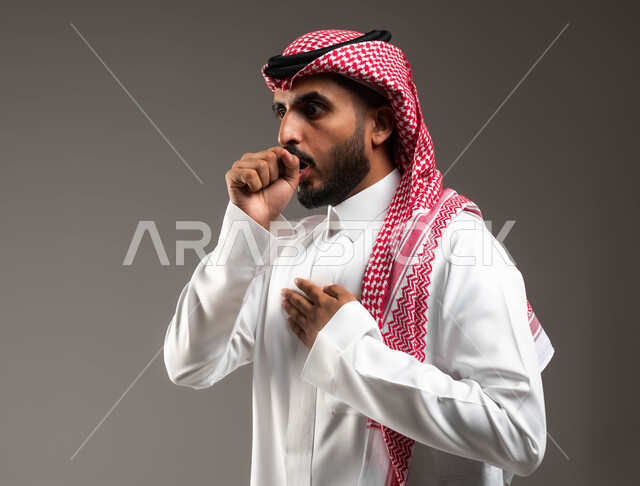 إيماءات الإرهاق والتعب الشديد، الإصابة بالأنفلونزا ونزلات البرد، الكحة والتهاب الحلق والحاجة للعلاج، بورتريه مقرب من الجانب لرجل عربي سعودي خليجي يرتدي الشماغ والثوب التقليدي ينظر إلى الكاميرا يضع يده أمام فمه ويقوم بالسعال,  خلفية رمادية .
